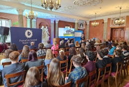 Szerokie ujęcie bogato zdobionej sali konferencyjnej wypełnionej słuchaczami siedzącymi w rzędach drewnianych krzeseł. Na środku, przed publicznością, stoi prelegent omawiający prezentację wyświetlaną na ekranie, która pokazuje tradycyjny drewniany dom. Wnętrze oświetlają klasyczne żyrandole, a sufit zdobią misterne sztukaterie.