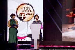 Elegancka gala wręczenia nagród z dużym podświetlonym logo „Złoty Medal 2026” na ekranie w tle. Dwie postacie stoją na scenie – jedna z notesem po lewej stronie, druga za przezroczystą mównicą po prawej. Oświetlenie sceniczne w odcieniach różu i fioletu podkreśla uroczysty charakter wydarzenia. W tle widoczny fragment aranżacji wnętrza wyświetlony na dużym ekranie.