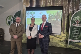 Trójka prelegentów stoi na scenie podczas konferencji w Technikum Leśnym w Tucholi, pozując na tle ekranu multimedialnego i leśnej grafiki. Po lewej stronie znajduje się mężczyzna w oliwkowym mundurze leśnika, w środku kobieta w eleganckim białym żakiecie, a po prawej mężczyzna w granatowym garniturze w kratę z czerwoną krawatką. Prezentacja wyświetlana w tle dotyczy edukacji w zmieniającym się klimacie, a po bokach widoczne są flagi reklamowe Branżowego Centrum Umiejętności.