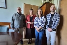 Grupa czterech osób, dwóch mężczyzn i dwie kobiety, stoi w biurowym pomieszczeniu o jasnych ścianach i drewnianej podłodze. Uczestnicy są ustawieni w rzędzie obok owalnego stołu konferencyjnego, a w tle widać wysoką szafę, na której wyeksponowano liczne puchary i trofea sportowe. Osoby ubrane są w stroje biznesowe oraz codzienne, w tym czerwoną marynarkę, wzorzystą koszulę i flanelową kratę. Wnętrze dopełniają tapicerowane drzwi oraz oprawiony dokument zawieszony na ścianie po lewej stronie.