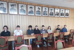 Grupa młodych ludzi siedzi przy długim, drewnianym stole konferencyjnym w sali o ścianach wyłożonych jasną boazerią. Nad ich głowami wisi rząd jednakowych, czarno-białych portretów przedstawiających mężczyzn w togach i nakryciach głowy, prawdopodobnie rektorów lub profesorów uczelni. Osoby siedzące przy stole są ubrane w codzienne stroje – t-shirty, bluzy i swetry – i patrzą przed siebie w stronę obiektywu.