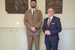 Dwóch mężczyzn stoi obok siebie na tle jasnej ściany, na której zawieszono dwie pamiątkowe, brązowe tablice z płaskorzeźbami. Postać po lewej stronie ma na sobie oliwkowy mundur leśnika, natomiast mężczyzna po prawej ubrany jest w elegancki, ciemnoniebieski garnitur i wzorzysty krawat. Obaj patrzą prosto w obiektyw z dłońmi splecionymi przed sobą, zachowując oficjalną postawę. Wnętrze jest nowoczesne i jasne, wykończone szklaną balustradą oraz beżowymi płytkami podłogowymi