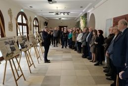 Mężczyzna w ciemnym garniturze przemawia do grupy osób zgromadzonych w jasnym, przestronnym korytarzu o kamiennej posadzce. Po lewej stronie, na drewnianych sztalugach, wyeksponowano tablice informacyjne ze zdjęciami i tekstami, które stanowią element wystawy historycznej. Słuchacze stoją w rzędzie wzdłuż ściany, uważnie obserwując prelegenta gestykulującego dłońmi.