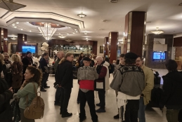 Tłum zgromadzony w przestronnym foyer o jasnej, marmurowej podłodze i podwieszanych, geometrycznych lampach sufitowych. Grupy osób rozmawiają w kilku punktach sali, część stoi, część siedzi w tle przy stolikach. Wnętrze ma elegancki charakter – dominują drewniane kolumny, lustra i ciepłe oświetlenie. Po prawej stronie znajduje się ekran informacyjny, a dalej widać balony oraz duże okno z widokiem na zewnętrzną część budynku. 