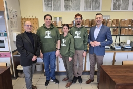 Pięciu pracowników naukowych stoi ramię w ramię w jasnej pracowni uniwersyteckiej, otoczonej próbkami drewna i sprzętem laboratoryjnym. Trzy osoby w centrum mają na sobie zielone bluzy z kapturem z logo „University of Forestry”, natomiast mężczyźni po bokach ubrani są w eleganckie marynarki. Tło wypełniają liczne eksponaty dendrologiczne ułożone na półkach oraz tablice z przekrojami pni drzew zawieszone na ścianie. Cała grupa uśmiecha się w stronę obiektywu.