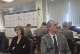 Uczestnicy spotkania siedzą przy długim, ciemnym stole w jasnej sali konferencyjnej, na której ścianie wisi czarny krzyż. W centrum uwagi znajduje się mężczyzna w okularach i kraciastej marynarce oraz kobieta z założonymi rękami, oboje patrzący z uwagą w tym samym kierunku. W tle widoczna jest ścianka reklamowa z logo Lasów Państwowych, przy której pracują inne osoby w oliwkowych mundurach leśnych. Na blacie przed siedzącymi stoją filiżanki z kawą oraz leżą akcesoria biurowe.