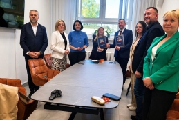Grupa osób stoi wokół długiego, szarego stołu w jasnym gabinecie. Dwoje uczestników w centrum trzyma pamiątkowe dyplomy lub teczki z godłem, symbolizując zakończenie oficjalnego spotkania lub podpisanie współpracy. Na pierwszym planie po prawej stronie wyróżnia się uśmiechnięta kobieta w jaskrawozielonej marynarce. Wnętrze dopełniają nowoczesne, brązowe fotele oraz duże okno, przez które wpada naturalne światło.