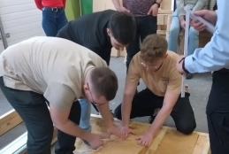 Młodzi mężczyźni, pochyleni nad drewnianą konstrukcją szkieletową, precyzyjnie układają w niej grube bloki jasnobrązowego materiału izolacyjnego. Ich pracy z bliska przygląda się stojący obok kolega, podczas gdy reszta grupy studentów obserwuje proces z dalszego planu warsztatu. Po prawej stronie widoczna jest postać trzymająca w dłoni długą miarę budowlaną, gotową do sprawdzenia wymiarów.