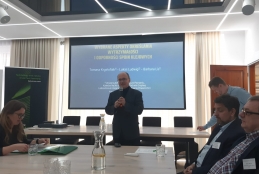 Mężczyzna w okularach i ciemnym garniturze przemawia do mikrofonu, stojąc przed dużym ekranem projekcyjnym z prezentacją naukową. Wokół długiego stołu konferencyjnego siedzą uczestnicy spotkania, w tym kobieta przeglądająca dokumenty po lewej stronie oraz dwaj mężczyźni na pierwszym planie po prawej. Na stole ustawione są szklane karafki z wodą, szklanki oraz materiały biurowe.