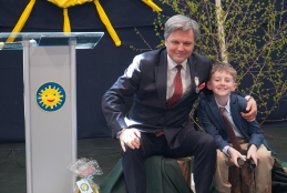 Uśmiechnięty mężczyzna w eleganckim garniturze i chłopiec w podobnym stroju siedzą na pieńkach na scenie. Mężczyzna obejmuje chłopca ramieniem, a w tle widać ozdobę w kształcie słońca oraz brzozowe gałązki. Obok stoi przezroczysta mównica z logiem żółtego słońca. Na podłodze przed nimi leży zafoliowany prezent, a na twarzach obu widać radość.