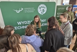 Grupa młodych osób stoi przy stoisku informacyjnym Uniwersytetu Przyrodniczego w Poznaniu podczas targów edukacyjnych. Uśmiechnięta przedstawicielka uczelni w zielonej koszulce rozmawia z zainteresowanymi, prezentując ofertę Wydziału Leśnego i Technologii Drewna. W tle widoczna jest duża zielona ścianka z logo uniwersytetu oraz materiały promocyjne rozłożone na blacie.