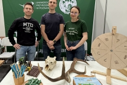 Trzej przedstawiciele Wydziału Leśnego i Technologii Drewna Uniwersytetu Przyrodniczego w Poznaniu stoją uśmiechnięci za stołem wystawowym. Na blacie przed nimi ułożono poroże, czaszkę zwierzęcia, drewniane koło fortuny oraz zielone gadżety i cukierki. Tło stanowi duża, zielona ścianka promocyjna z logo uczelni i nazwą wydziału. Całość tworzy profesjonalną, a zarazem przyjazną prezentację oferty edukacyjnej podczas wydarzenia targowego.