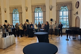 Kobieta przemawia do mikrofonu w eleganckiej, wysokiej sali z dużymi oknami i parkietem ułożonym w geometryczne wzory. Słuchacze stoją w skupieniu wokół wysokich stolików koktajlowych przykrytych czarnymi obrusami, a w tle widać czarny fortepian oraz bufet kawowy. Wnętrze oświetlają stylowe kinkiety, które podkreślają oficjalny, bankietowy charakter spotkania.