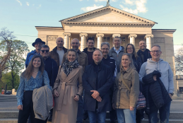 Grupa kilkunastu uśmiechniętych osób pozuje do wspólnej fotografii na szerokich, kamiennych schodach. W tle widoczny jest monumentalny budynek z klasycznymi kolumnami i trójkątnym frontonem, zwieńczony rzeźbą na dachu. Uczestnicy są ubrani w codzienne, wiosenne stroje, takie jak kurtki, płaszcze i swetry, a nad nimi rozciąga się jasne, błękitne niebo z delikatnymi chmurami. Całość kompozycji ma charakter pamiątkowego zdjęcia wykonanego przed zabytkowym obiektem w świetle dziennym.