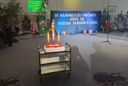 Na metalowym wózku gastronomicznym, ustawionym na środku hali sportowej, stoi prostokątny tort udekorowany owocami, z którego strzelają trzy płonące fontanny tortowe. W tle widoczna jest udekorowana scena z dużą, niebieską kurtyną opatrzoną białym napisem: „W NAJMNIEJSZEJ MRÓWCE KRYJE SIĘ WIELKA TAJEMNICA LASU”. Po bokach sceny zaaranżowano leśną dekorację z żywymi drzewkami, polskimi flagami oraz modelami dzikich zwierząt, w tym sarny i wilka. Na szarej podłodze hali, wokół wózka z tortem, widoczne są cien