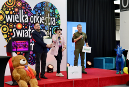 Trzy osoby stoją na scenie z czerwonym dywanem podczas finału Wielkiej Orkiestry Świątecznej Pomocy, co podkreśla wielkie, kolorowe serce w tle. Mężczyzna po lewej trzyma białą donicę z sadzonką drzewa, kobieta w różowej marynarce mówi do mikrofonu, a po prawej stoi mężczyzna w oliwkowej koszulce, trzymając dyplom.