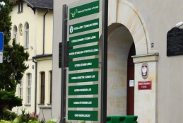 Przed wejściem do budynku Uniwersytetu Przyrodniczego w Poznaniu stoi wysoki, przeszklony pylon informacyjny z zielonymi tabliczkami. Wskazują one drogę do Wydziału Leśnego i Technologii Drewna oraz poszczególnych katedr, takich jak Katedra Inżynierii Leśnej czy Ogród Dendrologiczny. W tle widoczna jest jasna elewacja gmachu z klasycznym, łukowatym portalem wejściowym, obok którego umieszczono godło Polski i tablice pamiątkowe. Całość otacza zadbana zieleń ogrodowa i fragmenty zabytkowej architektury z char