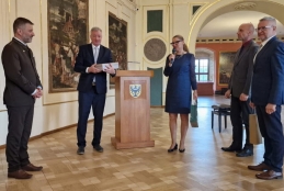 Grupa pięciu osób uczestniczy w oficjalnym spotkaniu w eleganckiej sali bankietowej zdobionej klasycznymi obrazami. W centralnym punkcie kobieta przemawia do mikrofonu, stojąc obok drewnianej mównicy z herbem przedstawiającym czarnego orła. Pozostali uczestnicy, ubrani w garnitury, obserwują wystąpienie, a jeden z mężczyzn trzyma w dłoniach płaskie, ozdobne pudełko. W tle widoczne są wysokie arkady prowadzące do kolejnych pomieszczeń oraz fragment czarnego fortepianu.
