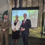 Trójka prelegentów stoi na scenie podczas konferencji w Technikum Leśnym w Tucholi, pozując na tle ekranu multimedialnego i leśnej grafiki. Po lewej stronie znajduje się mężczyzna w oliwkowym mundurze leśnika, w środku kobieta w eleganckim białym żakiecie, a po prawej mężczyzna w granatowym garniturze w kratę z czerwoną krawatką. Prezentacja wyświetlana w tle dotyczy edukacji w zmieniającym się klimacie, a po bokach widoczne są flagi reklamowe Branżowego Centrum Umiejętności.