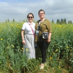 Dwie uśmiechnięte kobiety, prof. dr hab. Kinga Stuper-Szablewska oraz prof. UPP dr hab. Danuta Kurasiak-Popowska, stoją pośród wysokiej roślinności na kwitnącym polu. Jedna z nich ma na sobie białą koszulkę i wzorzystą spódnicę, a druga oliwkową koszulę i ciemne spodnie. W tle widać rozległą uprawę z żółtymi kwiatami pod lekko zachmurzonym niebem.