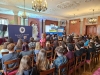 Szerokie ujęcie bogato zdobionej sali konferencyjnej wypełnionej słuchaczami siedzącymi w rzędach drewnianych krzeseł. Na środku, przed publicznością, stoi prelegent omawiający prezentację wyświetlaną na ekranie, która pokazuje tradycyjny drewniany dom. Wnętrze oświetlają klasyczne żyrandole, a sufit zdobią misterne sztukaterie.