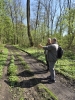 Dwóch mężczyzn stoi na polnej drodze pośród liściastego lasu w słoneczny dzień. Jeden z nich, ubrany w szary polar i plecak, wykonuje gest dłonią, jakby coś tłumaczył lub wskazywał kierunek swojemu towarzyszowi. Wokół drogi rośnie gęsta, zielona roślinność z drobnymi, żółtymi kwiatami, a wysokie drzewa o młodych liściach przepuszczają wyraźne smugi światła. Ścieżka nosi ślady kół pojazdów, tworząc ciemne, błotniste pasy na tle jasnej zieleni runa.