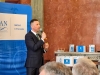 Mężczyzna w ciemnym garniturze stoi na tle bordowej ściany i mówi do mikrofonu trzymanego w prawej ręce. Obok niego znajduje się niebieska ścianka z logo PAN Oddział w Poznaniu. W tle, na marmurowym kominku, widać materiały promocyjne festiwalu oraz białe pudełka z logo wydarzenia.