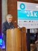 Starszy mężczyzna w okularach i ciemnym garniturze przemawia zza drewnianej mównicy, patrząc w dół na swoje notatki. Tuż za nim widoczny jest fragment planszy informacyjnej Poznańskiego Festiwalu Nauki i Sztuki 2026. Sceneria wskazuje na oficjalne otwarcie lub panel dyskusyjny w reprezentacyjnej sali.
