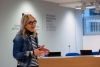 Kobieta w okularach i dżinsowej kurtce dynamicznie przemawia, gestykulując dłońmi. Stoi w nowoczesnej sali konferencyjnej przed drewnianym pulpitem, a w tle widać szarą ścianę z logotypami uczelni wyższej. Jej swobodny ubiór, składający się z pasiastej bluzki i czarnego golfa, kontrastuje z oficjalnym charakterem wnętrza. Całość kompozycji skupia uwagę na prelegentce uchwyconej w trakcie ożywionej wypowiedzi.