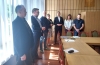 Zasłaniające okno żaluzje pionowe rzucają paski światła i cienia na grupę siedmiu osób zgromadzonych w sali konferencyjnej. Stoją oni w rzędzie wzdłuż ściany wyłożonej pionową boazerią drewnianą. Wnętrze dopełnia długi, drewniany stół z ułożonymi na nim dokumentami oraz ustawione wokół niego krzesła z oparciami wyściełanymi czerwoną i zieloną tkaniną.