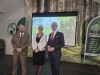 Trójka prelegentów stoi na scenie podczas konferencji w Technikum Leśnym w Tucholi, pozując na tle ekranu multimedialnego i leśnej grafiki. Po lewej stronie znajduje się mężczyzna w oliwkowym mundurze leśnika, w środku kobieta w eleganckim białym żakiecie, a po prawej mężczyzna w granatowym garniturze w kratę z czerwoną krawatką. Prezentacja wyświetlana w tle dotyczy edukacji w zmieniającym się klimacie, a po bokach widoczne są flagi reklamowe Branżowego Centrum Umiejętności.