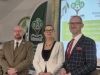 Trójka prelegentów pozuje do pamiątkowego zdjęcia podczas konferencji naukowej dotyczącej edukacji i leśnictwa. W centrum stoi kobieta w białym żakiecie i eleganckim naszyjniku, mając po lewej stronie mężczyznę w tradycyjnym, oliwkowym mundurze leśnika, a po prawej mężczyznę w granatowej marynarce w kratę z wyrazistym czerwonym krawatem. W tle widoczny jest fragment ekranu z logotypami BCU oraz zielona flaga reklamowa technikum.