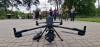 Duży, profesjonalny dron przemysłowy z zamontowaną pod spodem kamerą spoczywa na kostce brukowej w centralnej części kadru. Obok urządzenia leży aparatura sterująca, a w tle widać grupę osób stojących na trawniku i obserwujących pokaz. Scena rozgrywa się na otwartej przestrzeni w otoczeniu wysokich drzew i nowoczesnej architektury przy pochmurnym niebie. Perspektywa z niskiego poziomu podkreśla rozmiar i zaawansowaną konstrukcję bezzałogowca.