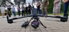 Duży, profesjonalny dron przemysłowy z zamontowaną pod spodem kamerą spoczywa na kostce brukowej w centralnej części kadru. Obok urządzenia leży aparatura sterująca, a w tle widać grupę osób stojących na trawniku i obserwujących pokaz. Scena rozgrywa się na otwartej przestrzeni w otoczeniu wysokich drzew i nowoczesnej architektury przy pochmurnym niebie. Perspektywa z niskiego poziomu podkreśla rozmiar i zaawansowaną konstrukcję bezzałogowca.
