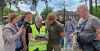 Kobieta w jaskrawej kamizelce odblaskowej prezentuje grupie osób obsługę profesjonalnego kontrolera do drona. Uczestnicy pokazu z bliska przyglądają się ekranowi urządzenia, podczas gdy jedna z osób dokumentuje proces telefonem. Spotkanie odbywa się na zewnątrz, na wybrukowanym placu w otoczeniu drzew i niskiej zabudowy. W tle widać pozostałych obserwatorów, którzy z zaciekawieniem śledzą przebieg warsztatów technologicznych.