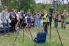 Grupa młodych ludzi stoi na trawiastym terenie, przyglądając się prezentacji profesjonalnego sprzętu pomiarowego ustawionego na statywach. Kobieta w jaskrawej, odblaskowej kamizelce obsługuje tablet, tłumacząc zgromadzonym zasady działania skanera laserowego. W tle widoczne są zaparkowane samochody oraz ściana zielonych drzew, co sugeruje warsztaty lub pokaz technologiczny na świeżym powietrzu. Obok urządzeń na ziemi leży otwarta, niebieska walizka transportowa chroniąca precyzyjny sprzęt.