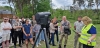 Grupa młodych ludzi stoi na trawiastym terenie, przyglądając się prezentacji profesjonalnego skanera laserowego ustawionego na statywie. Kobieta w jaskrawej, odblaskowej kamizelce obsługuje tablet, tłumacząc zgromadzonym zasady działania urządzenia marki FARO. W tle widać gęstą zieleń drzew oraz fragmenty nowoczesnej zabudowy, co sugeruje techniczne warsztaty plenerowe. Uczestnicy z zaciekawieniem obserwują pokaz, skupiając uwagę na nowoczesnym sprzęcie pomiarowym.