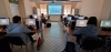 Uczniowie siedzą przy biurkach w sali komputerowej, pracując na stacjach roboczych wyposażonych w duże monitory. Na centralnej ścianie znajduje się ekran projekcyjny, na którym wyświetlana jest prezentacja techniczna lub interfejs specjalistycznego oprogramowania. Pomieszczenie jest jasne, oświetlone naturalnym światłem wpadającym przez okna oraz symetrycznie rozmieszczonymi lampami sufitowymi. Całość tworzy uporządkowaną przestrzeń sprzyjającą nauce i realizacji projektów cyfrowych.