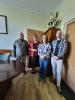 Grupa czterech osób, dwóch mężczyzn i dwie kobiety, stoi w biurowym pomieszczeniu o jasnych ścianach i drewnianej podłodze. Uczestnicy są ustawieni w rzędzie obok owalnego stołu konferencyjnego, a w tle widać wysoką szafę, na której wyeksponowano liczne puchary i trofea sportowe. Osoby ubrane są w stroje biznesowe oraz codzienne, w tym czerwoną marynarkę, wzorzystą koszulę i flanelową kratę. Wnętrze dopełniają tapicerowane drzwi oraz oprawiony dokument zawieszony na ścianie po lewej stronie.