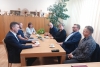 Grupa siedmiu osób siedzi naprzeciwko siebie przy prostokątnym, drewnianym stole w jasnym biurze. Na blacie rozstawione są filiżanki z kawą, talerzyk ze słodyczami oraz drobne akcesoria biurowe. W tle widoczne są wysokie, drewniane szafy z poukładanymi na nich pamiątkami i rękodziełem oraz duża roślina doniczkowa.