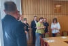 Grupa osób stoi w sali konferencyjnej wyłożonej pionową, drewnianą boazerią, skupiając uwagę na kobiecie w jasnozielonej bluzce i okularach. Kobieta trzyma w dłoniach drobny przedmiot, a obok niej stoi inna uczestniczka spotkania w białej koszuli. W tle widać pozostałych zebranych w strojach formalnych i codziennych, a na pierwszym planie po lewej stronie zarysowana jest sylwetka mężczyzny tyłem do obiektywu.