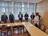 Grupa osób stoi w jasnej sali konferencyjnej, zgromadzona wokół połączonych w podkowę drewnianych stołów. Po prawej stronie wyróżnia się uśmiechnięta kobieta w białej koszuli w paski, która trzyma w dłoniach małą kartkę i zwraca się do pozostałych uczestników. W tle widoczne są duże okna przysłonięte pionowymi żaluzjami oraz ściana pokryta jasną, pionową boazerią. Wokół stołów, na których leżą pojedyncze dokumenty, ustawione są eleganckie krzesła z charakterystycznymi zielonymi obiciami.