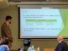 Mężczyzna w oliwkowym mundurze leśnika prowadzi prelekcję, stojąc obok dużego ekranu projekcyjnego z wyświetloną prezentacją. Na pierwszym planie widoczne są sylwetki słuchaczy oraz stół z butelkami wody mineralnej, co podkreśla konferencyjny charakter spotkania. Całość odbywa się w jasnej sali wykładowej, a uwaga osób w kadrze skupiona jest na prezentowanych danych.