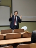 Mężczyzna w eleganckim ciemnym garniturze, białej koszuli i wzorzystym krawacie stoi przed audytorium w sali wykładowej. Energicznie gestykuluje obiema dłońmi, prowadząc prelekcję lub wykład dla osób siedzących w drewnianych ławkach widocznych na pierwszym planie. W tle znajduje się biała tablica oraz duży ekran projekcyjny, na którym wyświetlony jest interfejs odtwarzacza wideo. Obok prelegenta, na biurku, stoi nowoczesny biały głośnik.