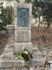 Kamienny obelisk z pamiątkową tablicą z brązu stoi w parku wśród drzew i krzewów. Tablica przedstawia profil Józefa Rivolego oraz poświęcony mu napis, a u podstawy pomnika leży wiązanka czerwonych kwiatów z zieloną wstęgą. Wokół monumentu widoczne są pozostałości śniegu oraz suche liście, co podkreśla zimową aurę.