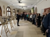 Mężczyzna w ciemnym garniturze przemawia do grupy osób zgromadzonych w jasnym, przestronnym korytarzu o kamiennej posadzce. Po lewej stronie, na drewnianych sztalugach, wyeksponowano tablice informacyjne ze zdjęciami i tekstami, które stanowią element wystawy historycznej. Słuchacze stoją w rzędzie wzdłuż ściany, uważnie obserwując prelegenta gestykulującego dłońmi.