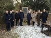 Grupa dziewięciu osób w zimowych płaszczach i kurtkach stoi w półkolu przed kamiennym pomnikiem Józefa Rivolego. Uczestnicy spotkania patrzą w stronę obiektywu, pozując do pamiątkowego zdjęcia w parkowej scenerii. Ziemia wokół nich jest lekko oszroniona i pokryta resztkami śniegu, a w tle widać gęste korony drzew i fragment jasnego budynku.