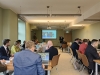 Uczestnicy konferencji siedzą przy długich stołach w jasnej, nowoczesnej sali, biorąc udział w spotkaniu hybrydowym. Na ekranie w głębi pomieszczenia wyświetlany jest widok z kamery przedstawiający kobietę uczestniczącą w dyskusji zdalnie, a obok ekranu stoi prelegent w ciemnym garniturze. Wiele osób korzysta z laptopów lub robi notatki, a na stołach widoczne są kubki z kawą i napoje, co sugeruje roboczy charakter wydarzenia.