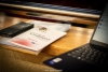 Program konferencji EU‑PoTaRCh leżący na drewnianej powierzchni obok smartfona i częściowo widocznego laptopa. Broszura ma biały front z logo projektu, pełnym tytułem wydarzenia i oznaczeniami partnerów. W tle widać metalową barierkę oraz miękkie, rozmyte oświetlenie sali, które nadaje scenie ciepły ton.