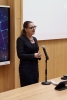 Kobieta w okularach i eleganckim, czarnym kostiumie stoi przy mikrofonie zamontowanym na statywie biurkowym. W tle widoczny jest fragment ekranu z nowoczesną, sieciową grafiką oraz gładka ściana o strukturze jasnego drewna. Prelegentka ma splecione dłonie i zdaje się odpowiadać na pytania lub prowadzić ożywioną prelekcję.